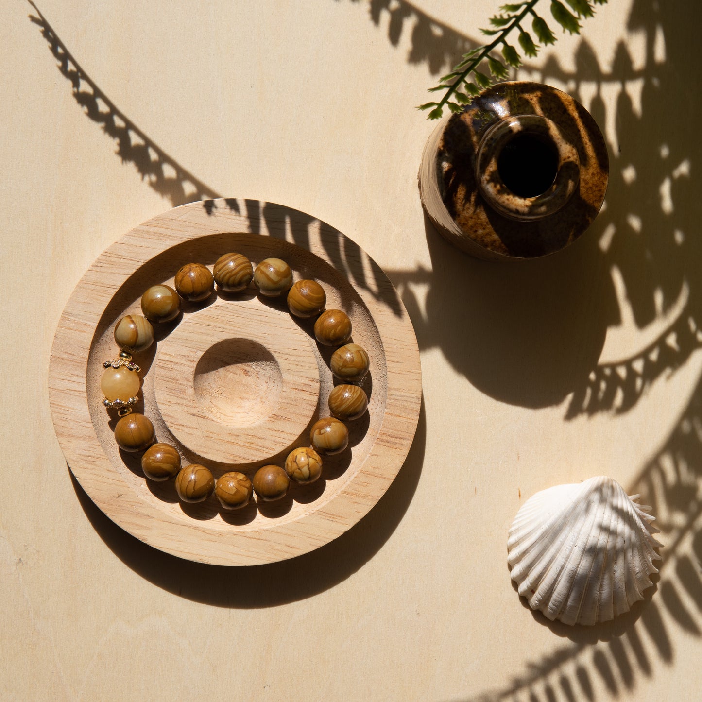 A lace wood bracelet sits on a wooden coaster, alongside a shell on a beige surface with plant shadows.