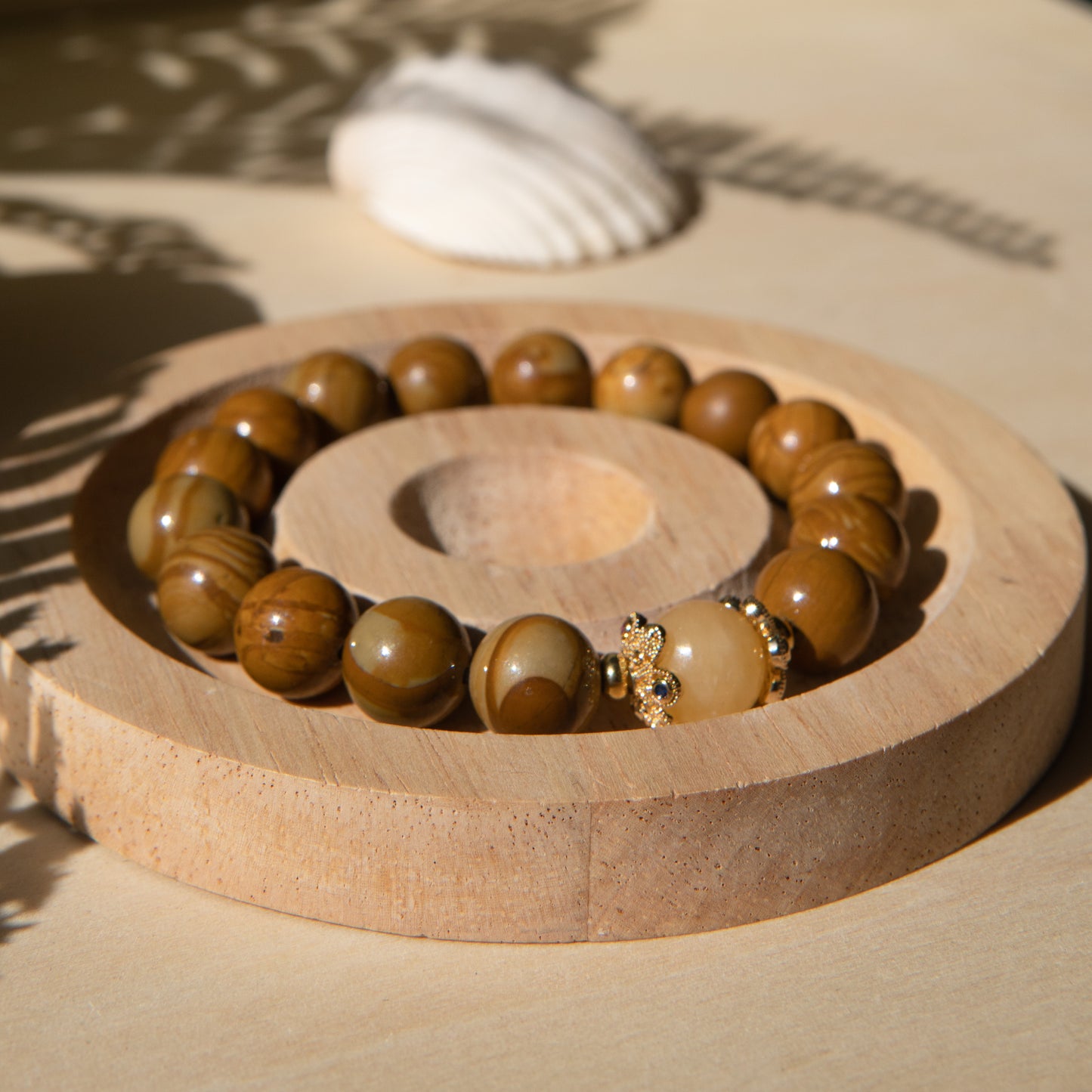 A lace wood bracelet sits on a wooden coaster, alongside a shell on a beige surface with plant shadows.
