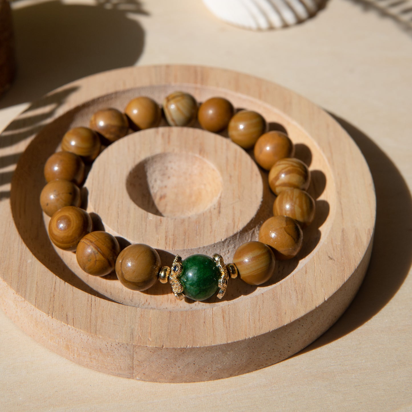 A lacewood beaded bracelet with a Malayian jade crystal, displayed on a wooden coaster with a shell in the background.