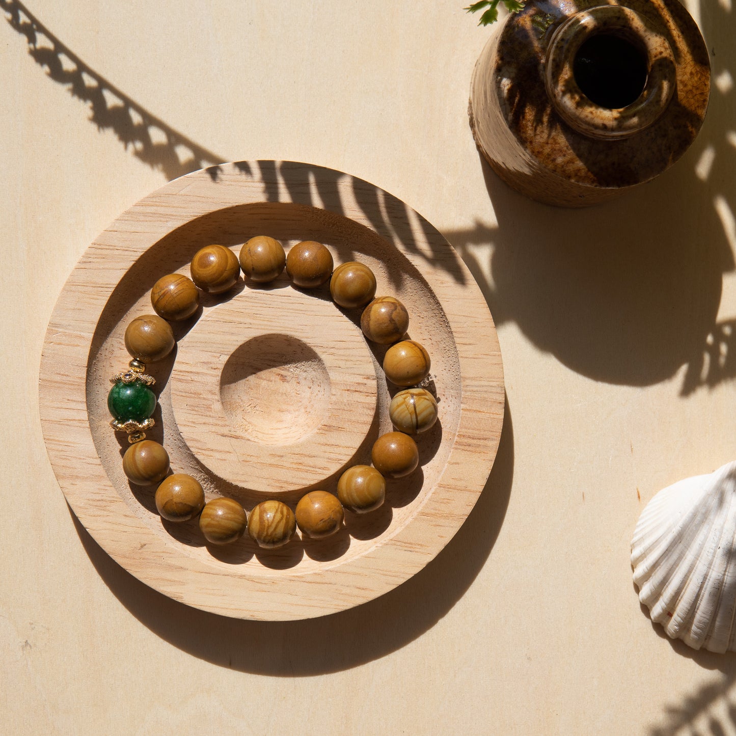 A lacewood beaded bracelet with a Malayian jade crystal, displayed on a wooden coaster with a shell in the background.