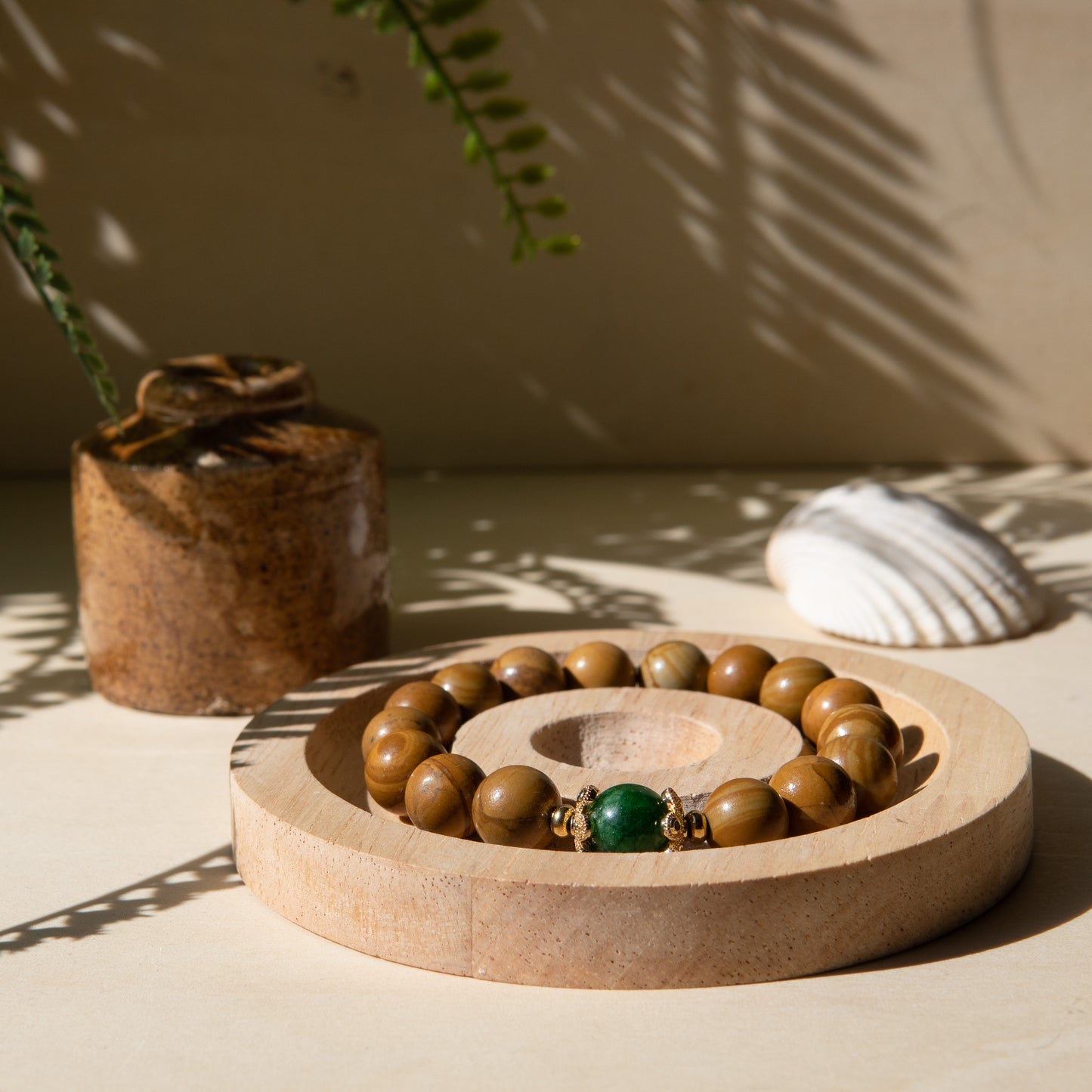 A lacewood beaded bracelet with a Malayian jade crystal, displayed on a wooden coaster with a shell in the background.
