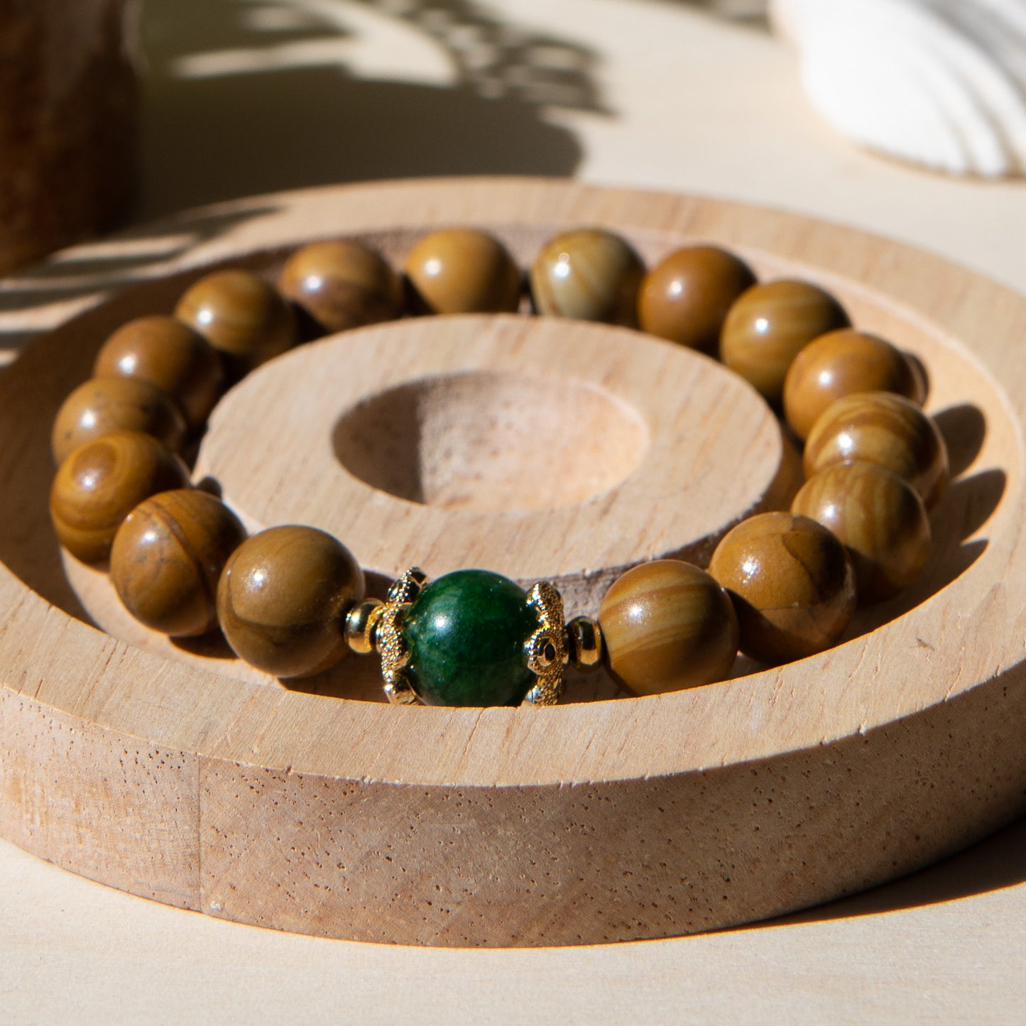 A lacewood beaded bracelet with a Malayian jade crystal, displayed on a wooden coaster with a shell in the background.