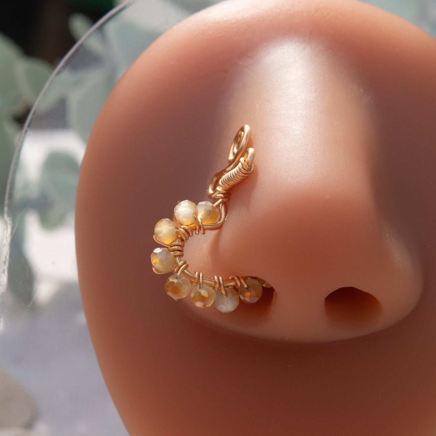 A gold-toned faux nose ring cuffs the model's nose. Beige transparent seed beads are wire-wrapped around the hoop's circumference. Above the hoop, the wire design forms an Ankh symbol.