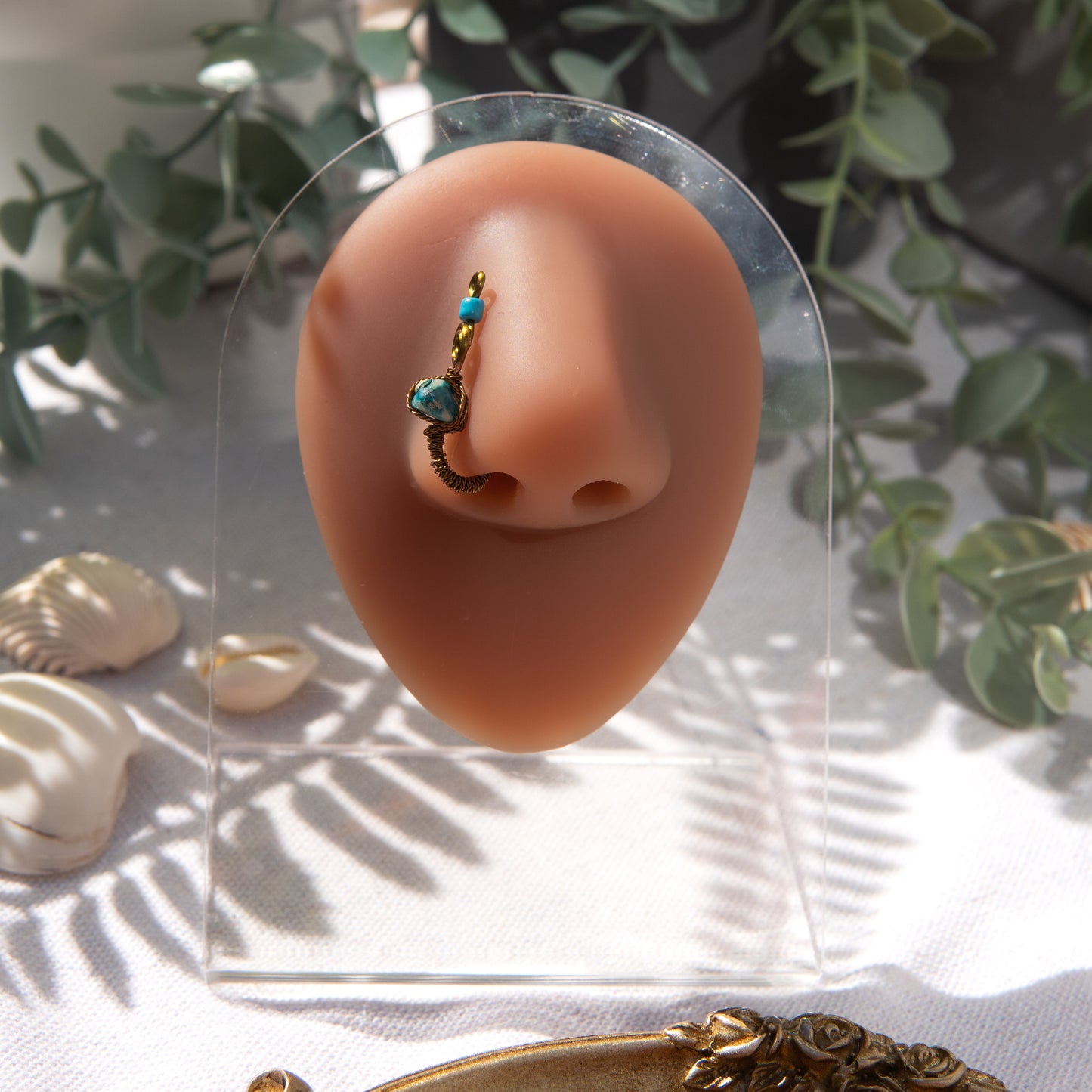 A bronze-toned faux nose ring cuffs the model's nostril. A turquoise crystal gemstone is wire-wrapped and interweaved into a vertical spiral wire design. A turquoise seed bead resides at the top of the cuff design.