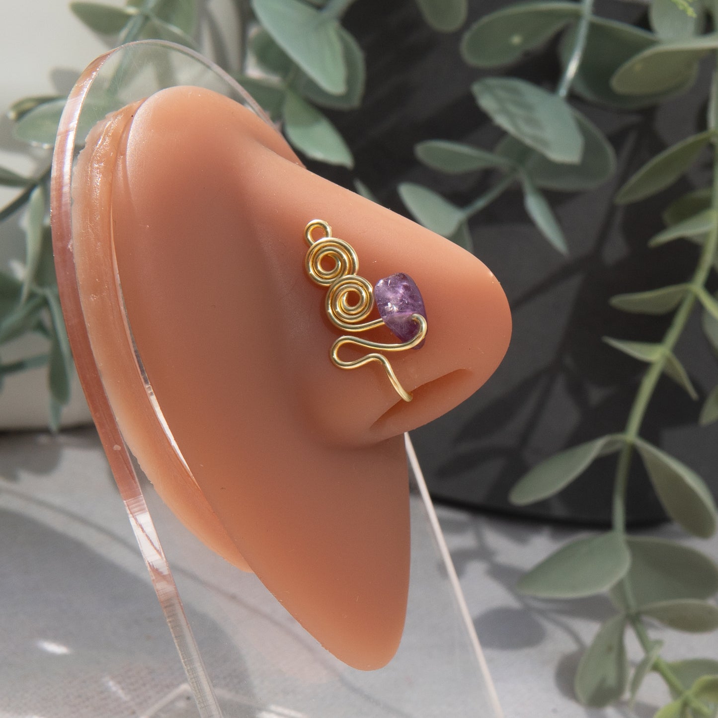 A gold-toned faux nose ring adorns the model's nose in a circular wire design that spirals up and gradually decreases in size. A crystal amethyst gemstone is woven into the design and rests at the bottom.