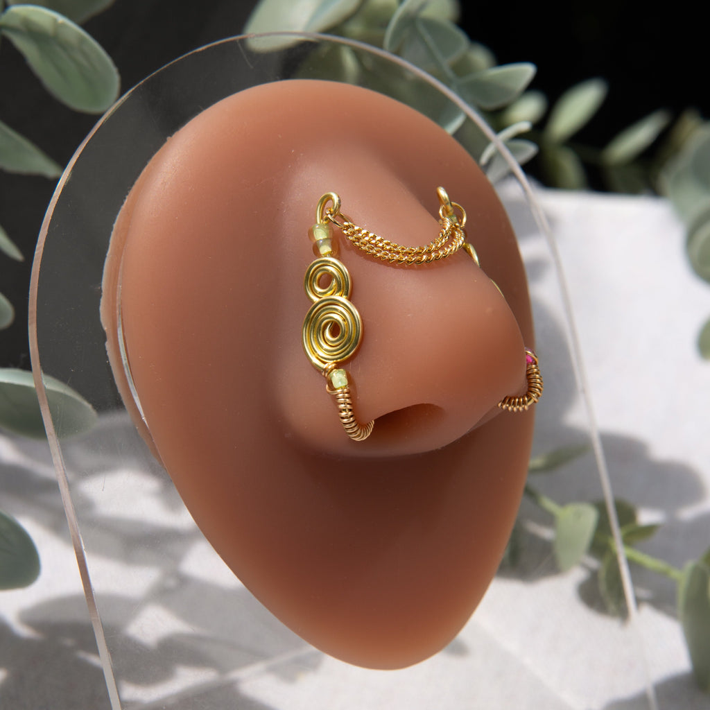 A two-toned faux bridge nose piercing in gold cuffs both nostrils of the model. Two dainty 18K gold-plated curb chains rest across the bridge of the nose, attached to the top of each nose cuff. Three seed beads are interweaved into the design of each nose cuff.