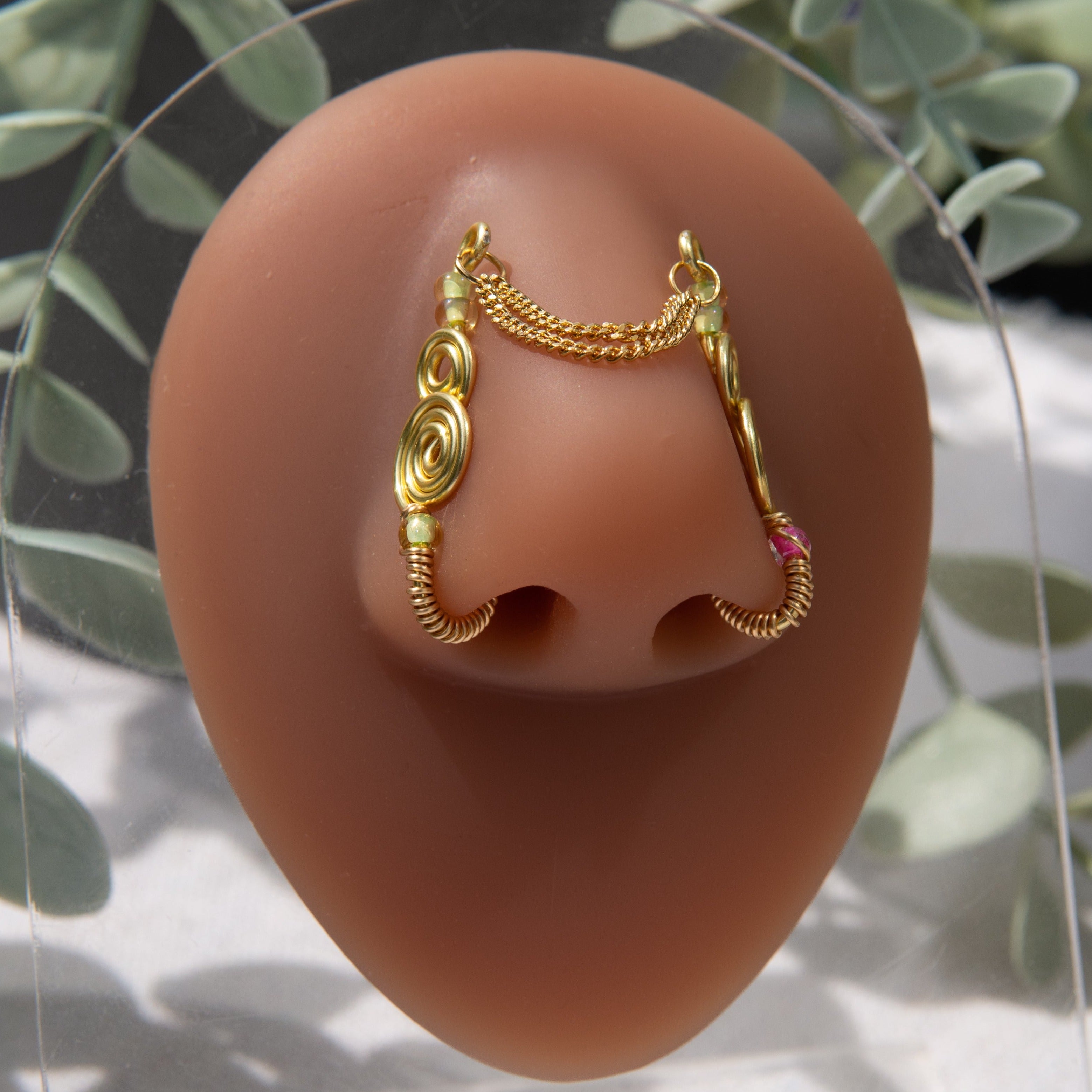 A two-toned faux bridge nose piercing in gold cuffs both nostrils of the model. Two dainty 18K gold-plated curb chains rest across the bridge of the nose, attached to the top of each nose cuff. Three seed beads are interweaved into the design of each nose cuff.