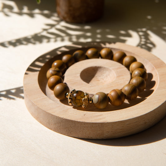 A lacewood beaded bracelet with a dragon viens agate crystal, displayed on a wooden coaster with a shell in the background.