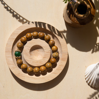 A lacewood beaded bracelet with a Malayian jade crystal, displayed on a wooden coaster with a shell in the background.