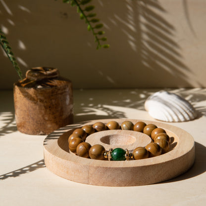 A lacewood beaded bracelet with a Malayian jade crystal, displayed on a wooden coaster with a shell in the background.