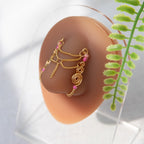 A gold-toned faux chain bridge nose piercing cuffs both of the model's nostrils. Two dainty 18K gold-plated curb chains rest across the bridge of the nose attached to the top and middle of each cuff, forming a criss-cross shape. Three pink seed beads were interweaved into the design of each nose cuff.