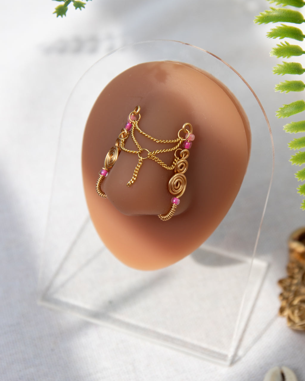 A gold-toned faux chain bridge nose piercing cuffs both of the model's nostrils. Two dainty 18K gold-plated curb chains rest across the bridge of the nose attached to the top and middle of each cuff, forming a criss-cross shape. Three pink seed beads were interweaved into the design of each nose cuff.