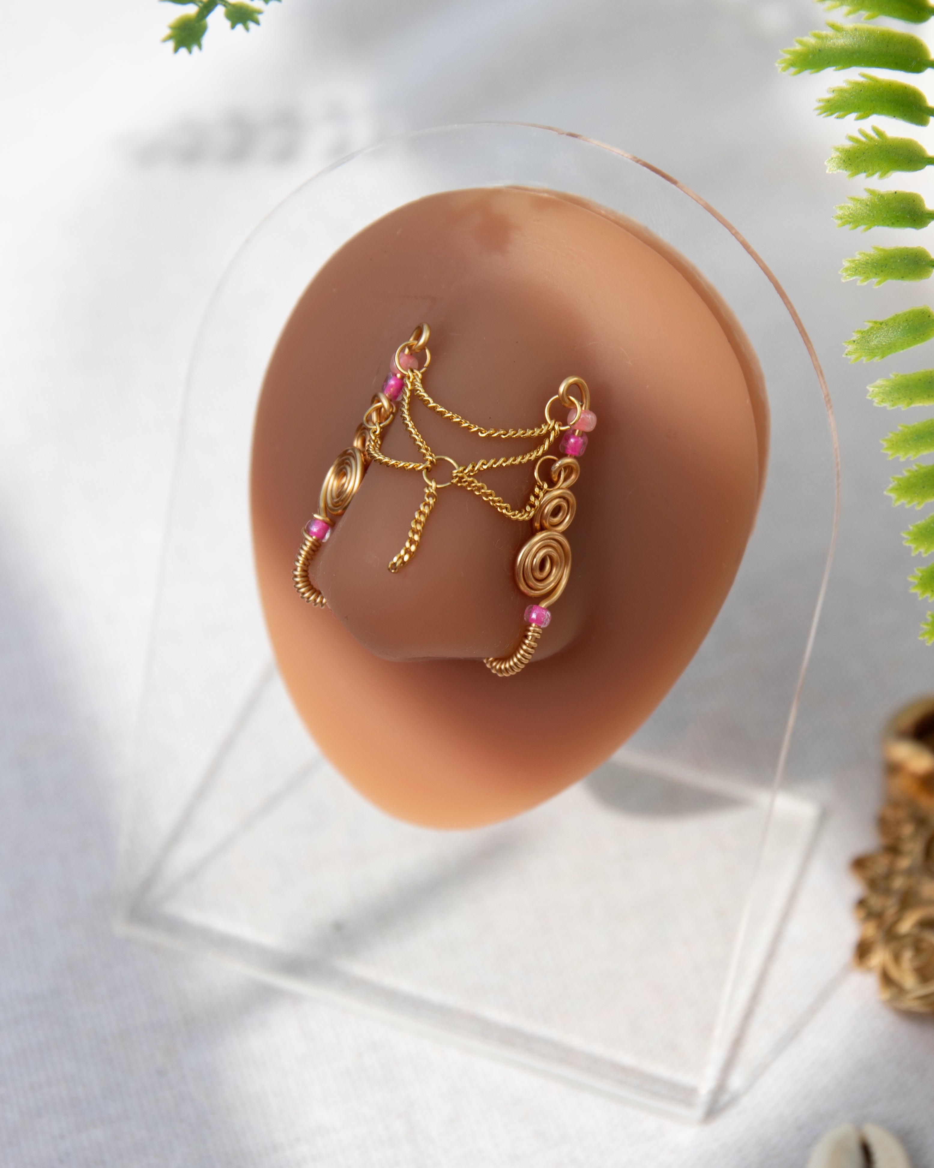 A gold-toned faux chain bridge nose piercing cuffs both of the model's nostrils. Two dainty 18K gold-plated curb chains rest across the bridge of the nose attached to the top and middle of each cuff, forming a criss-cross shape. Three pink seed beads were interweaved into the design of each nose cuff.