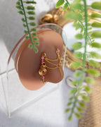 A gold-toned faux chain bridge nose piercing cuffs both of the model's nostrils. Two dainty 18K gold-plated curb chains rest across the bridge of the nose attached to the top and middle of each cuff, forming a criss-cross shape. Three pink seed beads were interweaved into the design of each nose cuff.