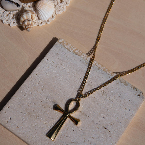 Gold-tone ankh pendant necklace on a matching chain, styled on a textured stone slab with seashells in the background.