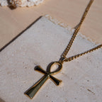 A gold-toned necklace with an ankh pendant resting on a textured light stone surface.