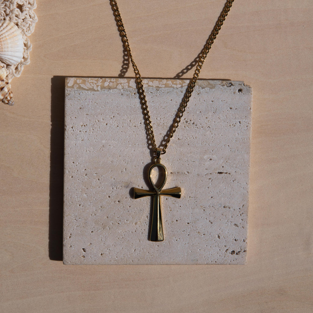A gold-toned chain necklace with an ankh pendant displayed on a square textured stone tile against a light wood background.