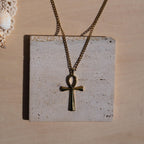A gold-toned chain necklace with an ankh pendant displayed on a square textured stone tile against a light wood background.