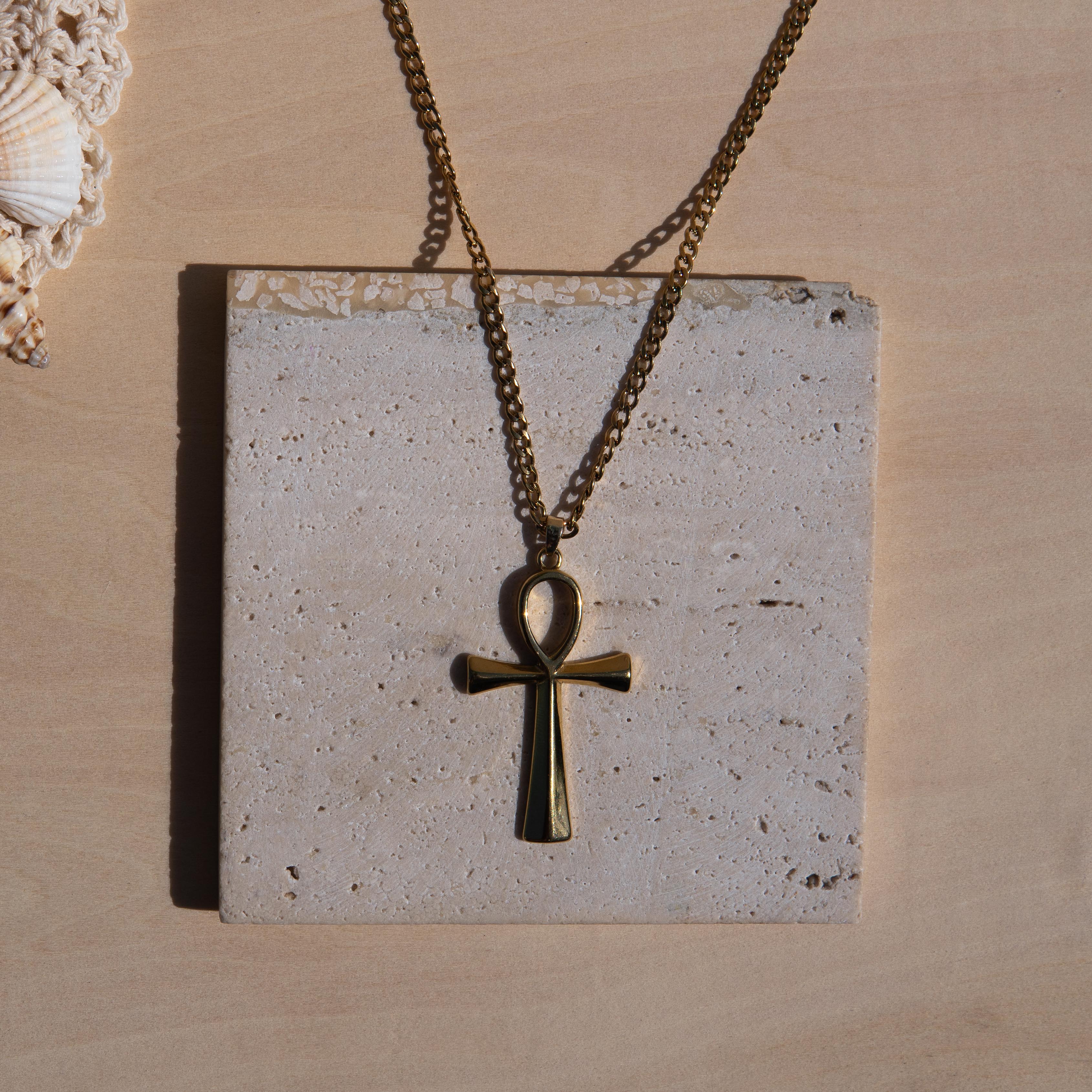 A gold-toned chain necklace with an ankh pendant displayed on a square textured stone tile against a light wood background.