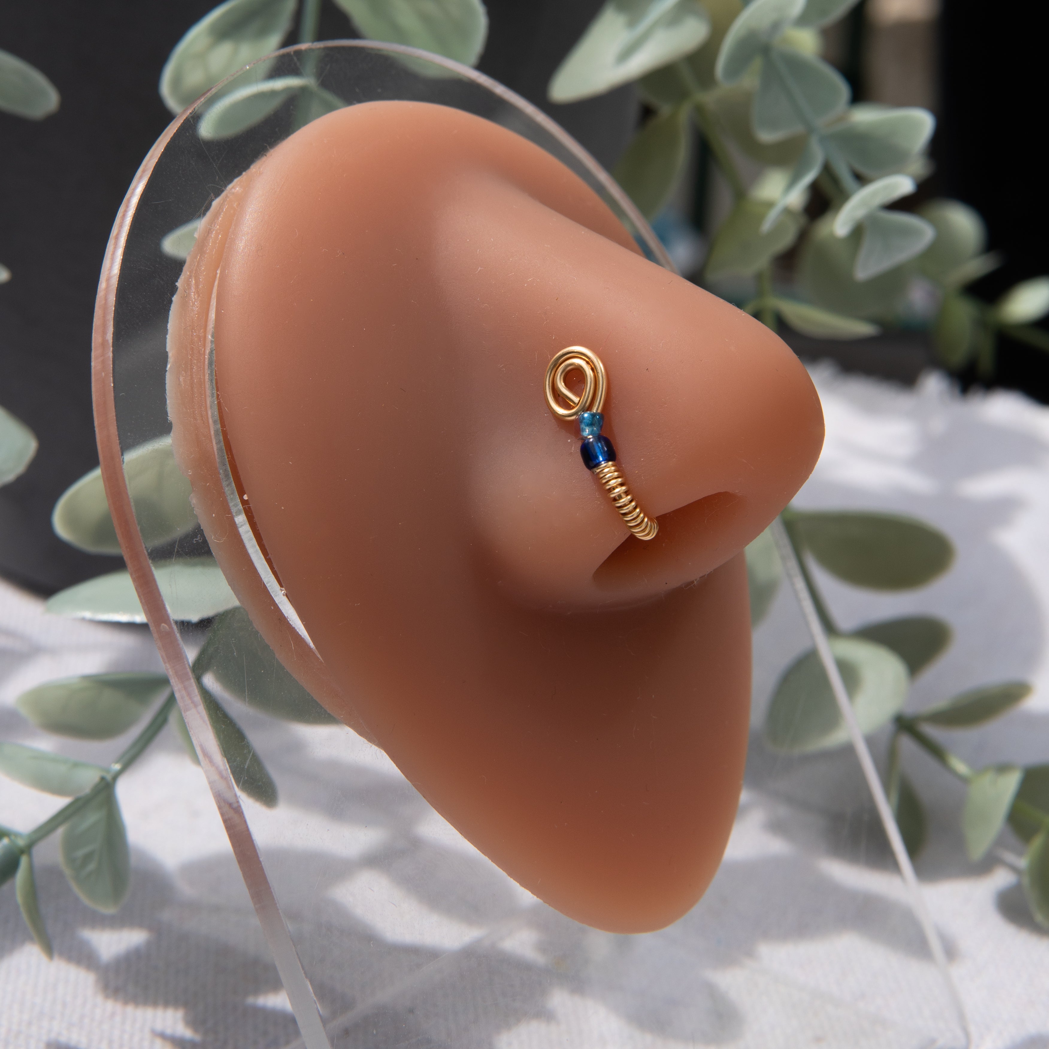 A gold-toned fake nose ring cuffs the model's nostril. The wire design forms a circular shape. Two blue seed beads rest below the circle.