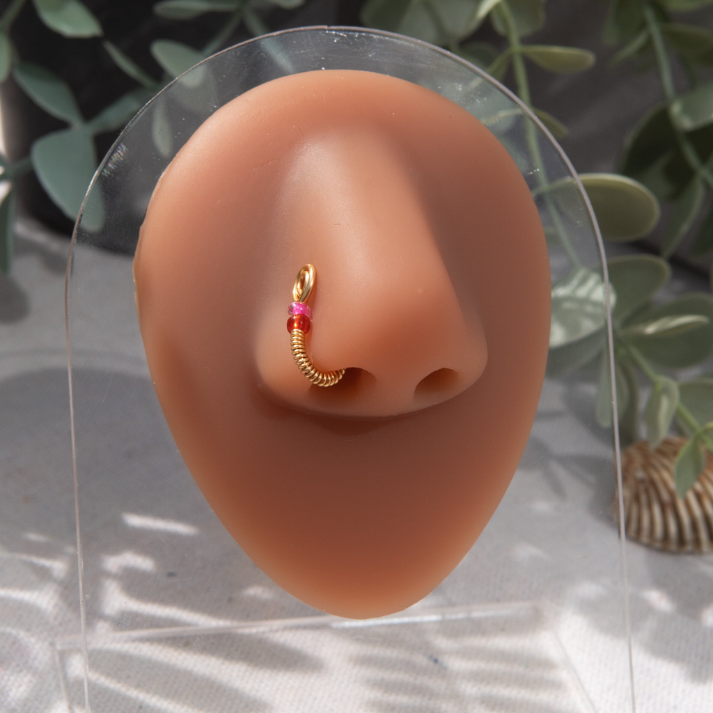 A gold-toned nose cuff rests on the model's nostril. The wire design forms a circular shape, with two pink seed beads resting below the circle.