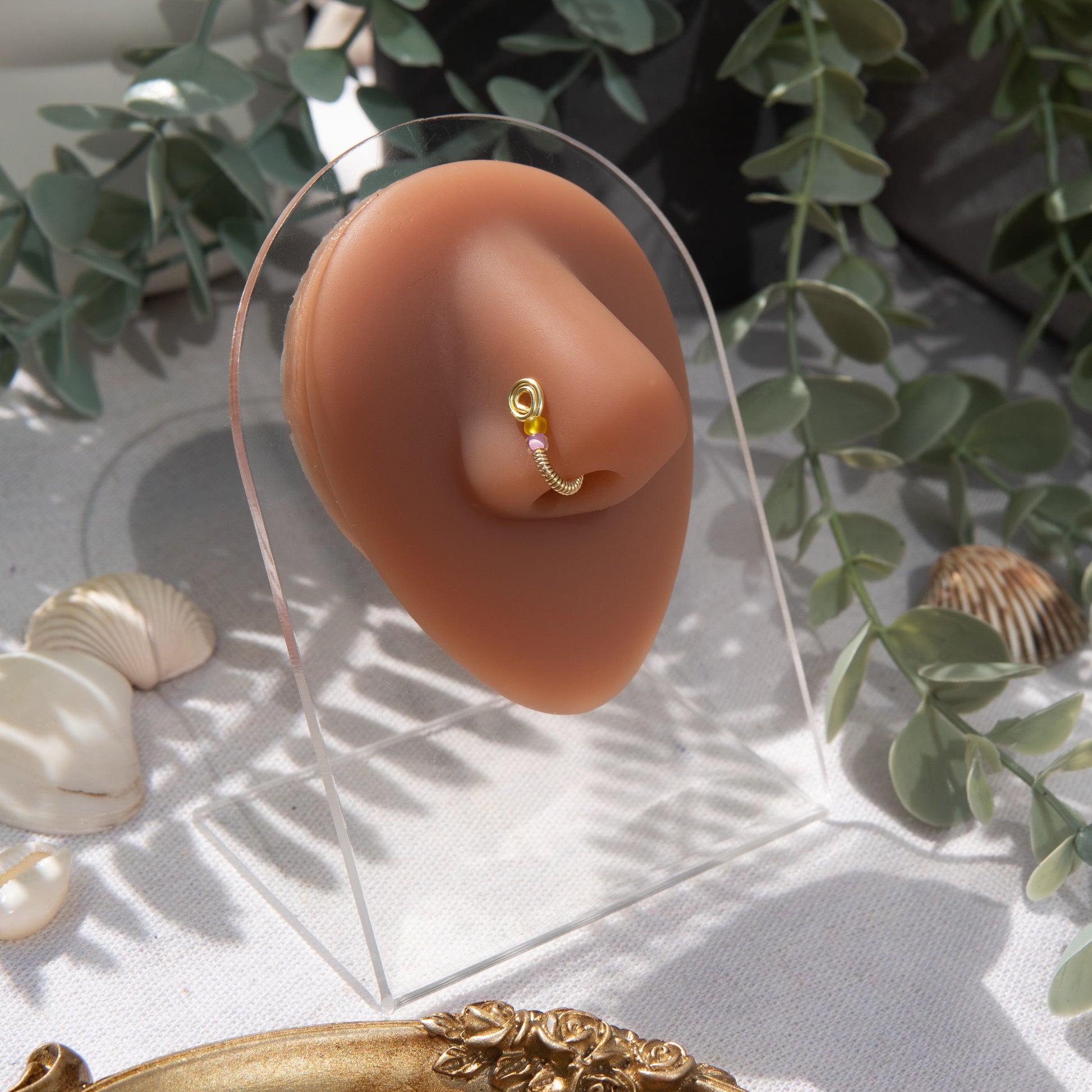 A light gold-toned fake nose ring cuffs the model's nostril. The wire design forms a circular shape. Two seed beads (yellow and lilac) rest below the circle.