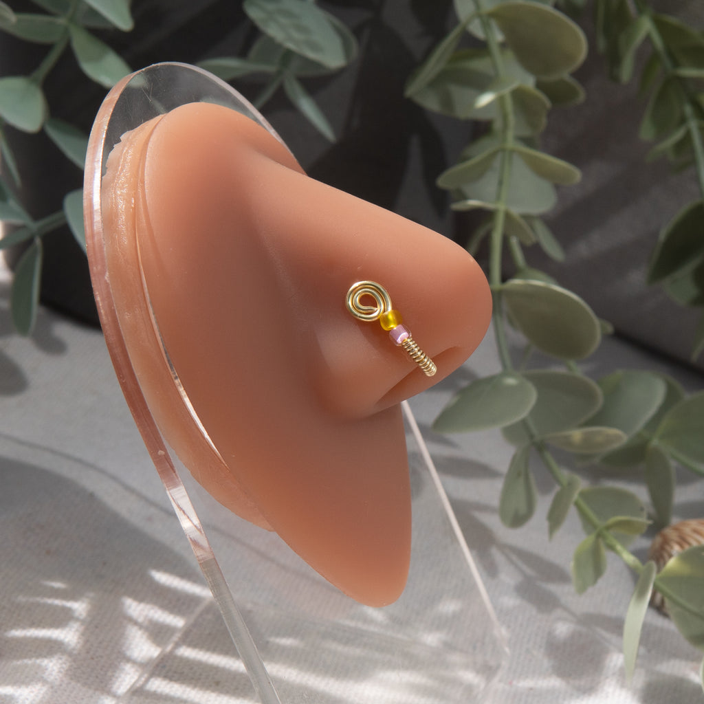 A light gold-toned fake nose ring cuffs the model's nostril. The wire design forms a circular shape. Two seed beads (yellow and lilac) rest below the circle.
