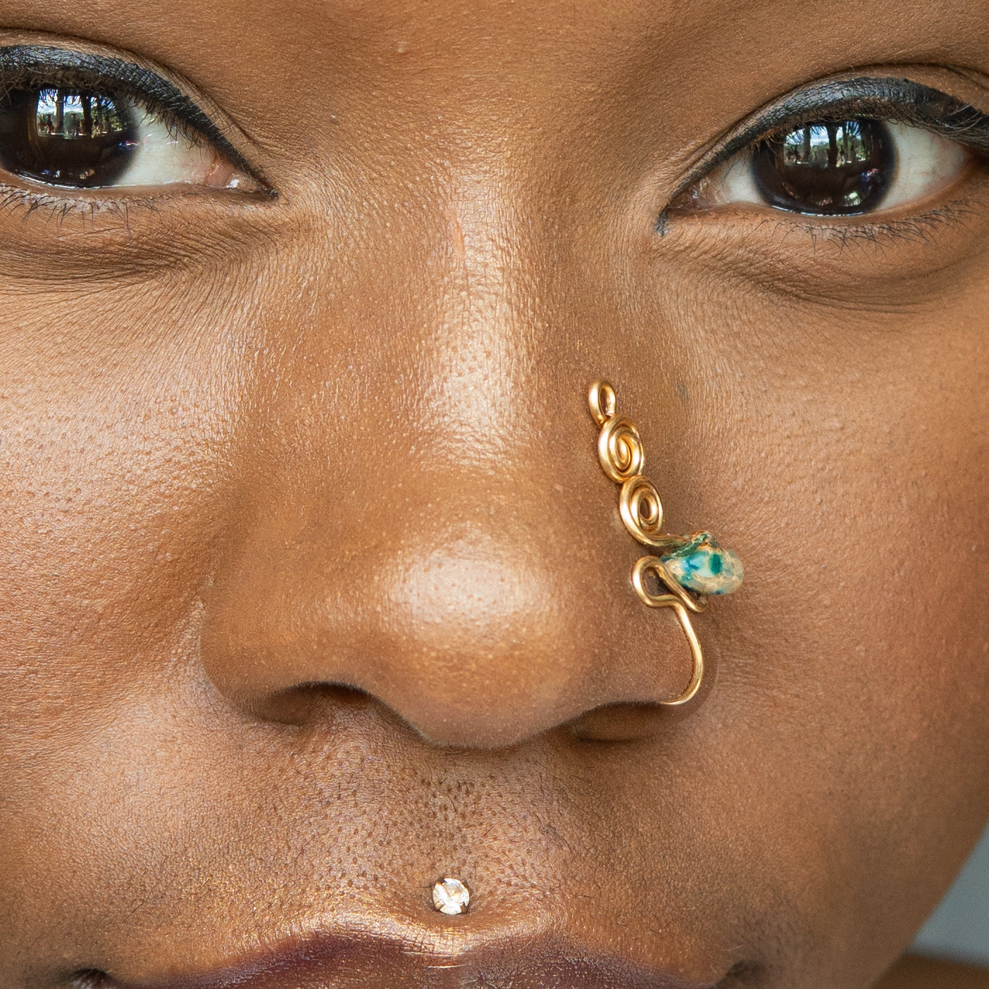 A gold-toned faux nose ring adorns the model's nose in a circular wire design that spirals up and gradually decreases in size. A crystal amazonite gemstone is woven into the design and rests at the bottom.