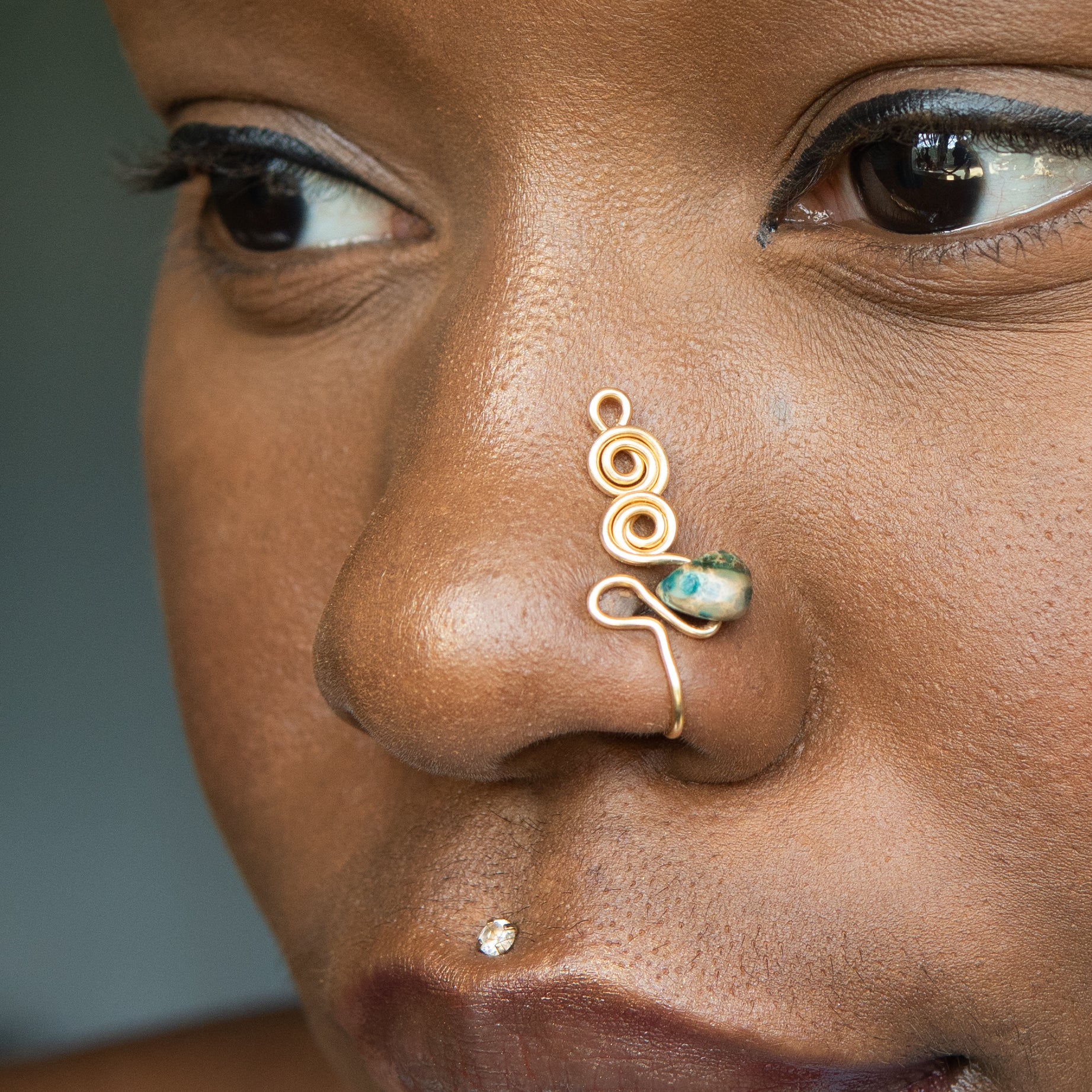 A gold-toned faux nose ring adorns the model's nose in a circular wire design that spirals up and gradually decreases in size. A crystal amazonite gemstone is woven into the design and rests at the bottom.