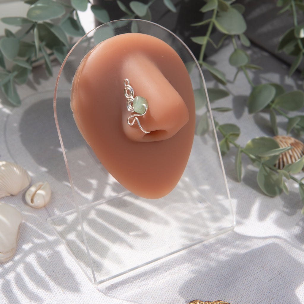 A silver-toned faux nose ring adorns the model's nose in a circular wire design that spirals up and gradually decreases in size. A crystal gemstone is woven into the design and rests at the bottom.