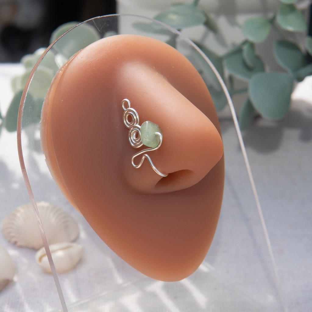 A silver-toned faux nose ring adorns the model's nose in a circular wire design that spirals up and gradually decreases in size. A crystal gemstone is woven into the design and rests at the bottom.