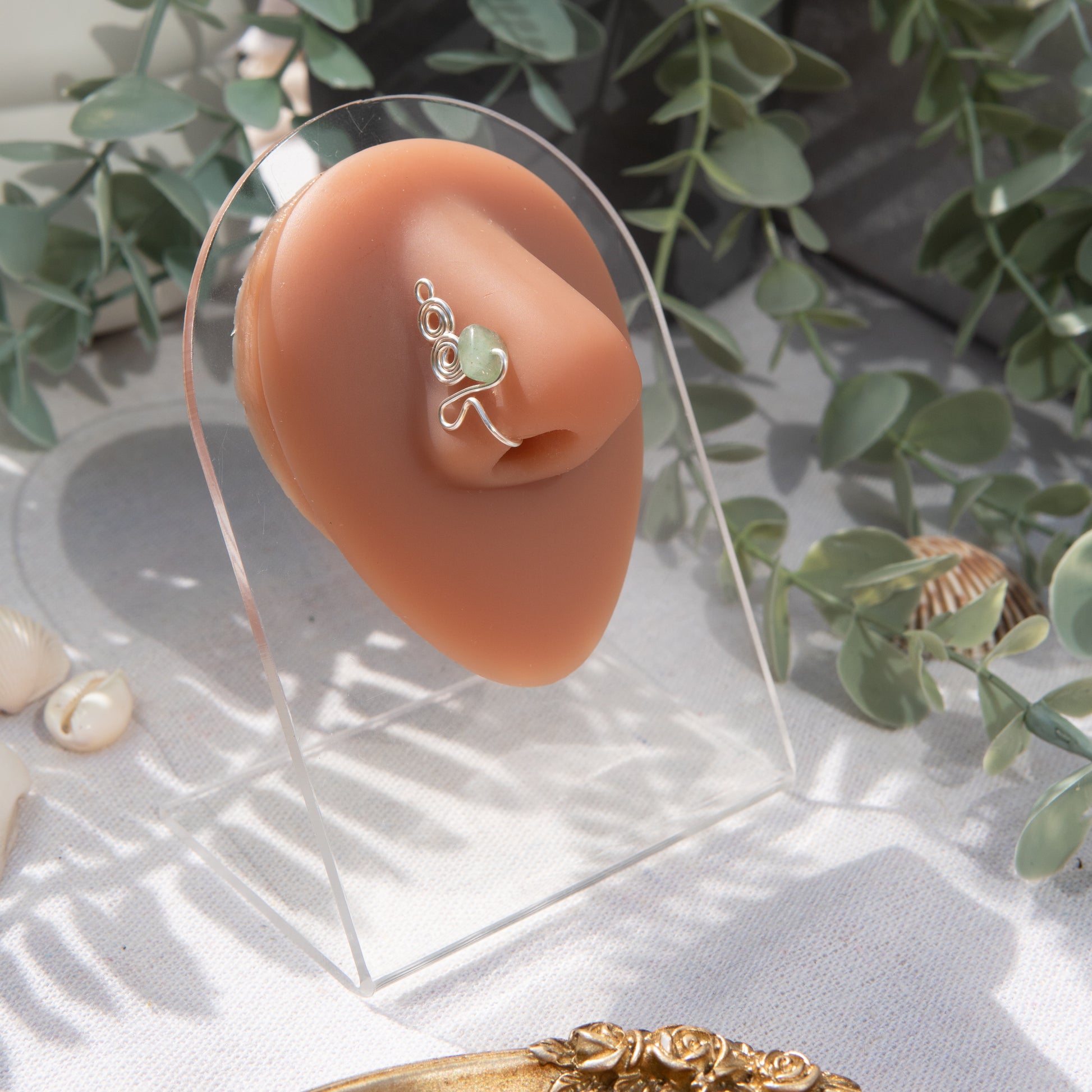A silver-toned faux nose ring adorns the model's nose in a circular wire design that spirals up and gradually decreases in size. A crystal gemstone is woven into the design and rests at the bottom.