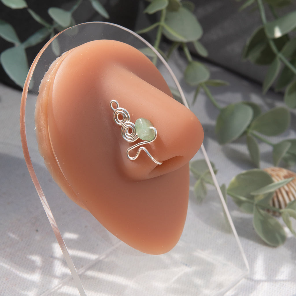 A silver-toned faux nose ring adorns the model's nose in a circular wire design that spirals up and gradually decreases in size. A crystal gemstone is woven into the design and rests at the bottom.