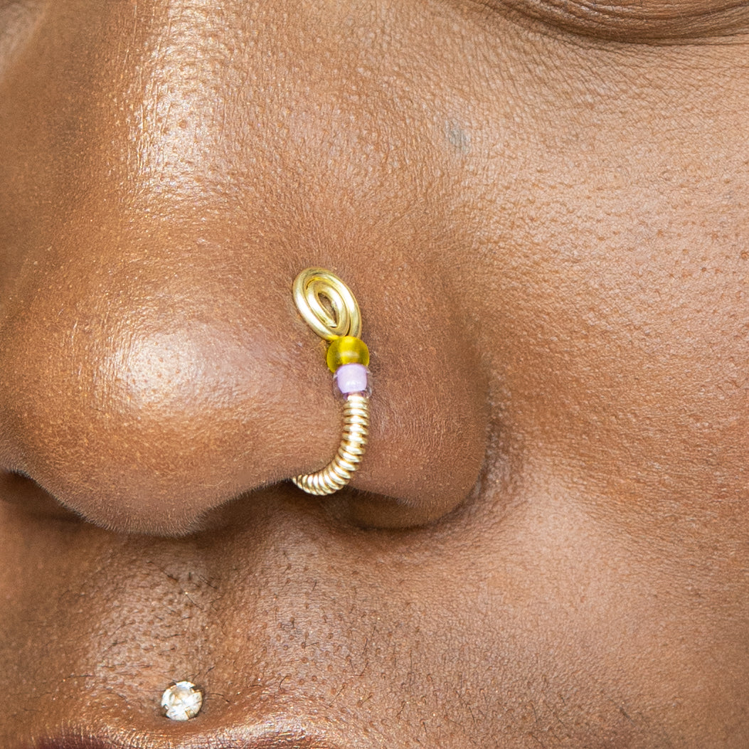 A light gold-toned fake nose ring cuffs the model's nostril. The wire design forms a circular shape. Two seed beads (yellow and lilac) rest below the circle.