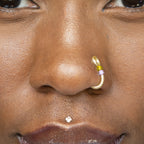 A light gold-toned fake nose ring cuffs the model's nostril. The wire design forms a circular shape. Two seed beads (yellow and lilac) rest below the circle.