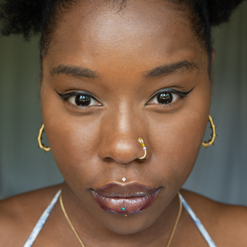 A light gold-toned fake nose ring cuffs the model's nostril. The wire design forms a circular shape. Two seed beads (yellow and lilac) rest below the circle.