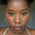 A light gold-toned fake nose ring cuffs the model's nostril. The wire design forms a circular shape. Two seed beads (yellow and lilac) rest below the circle.