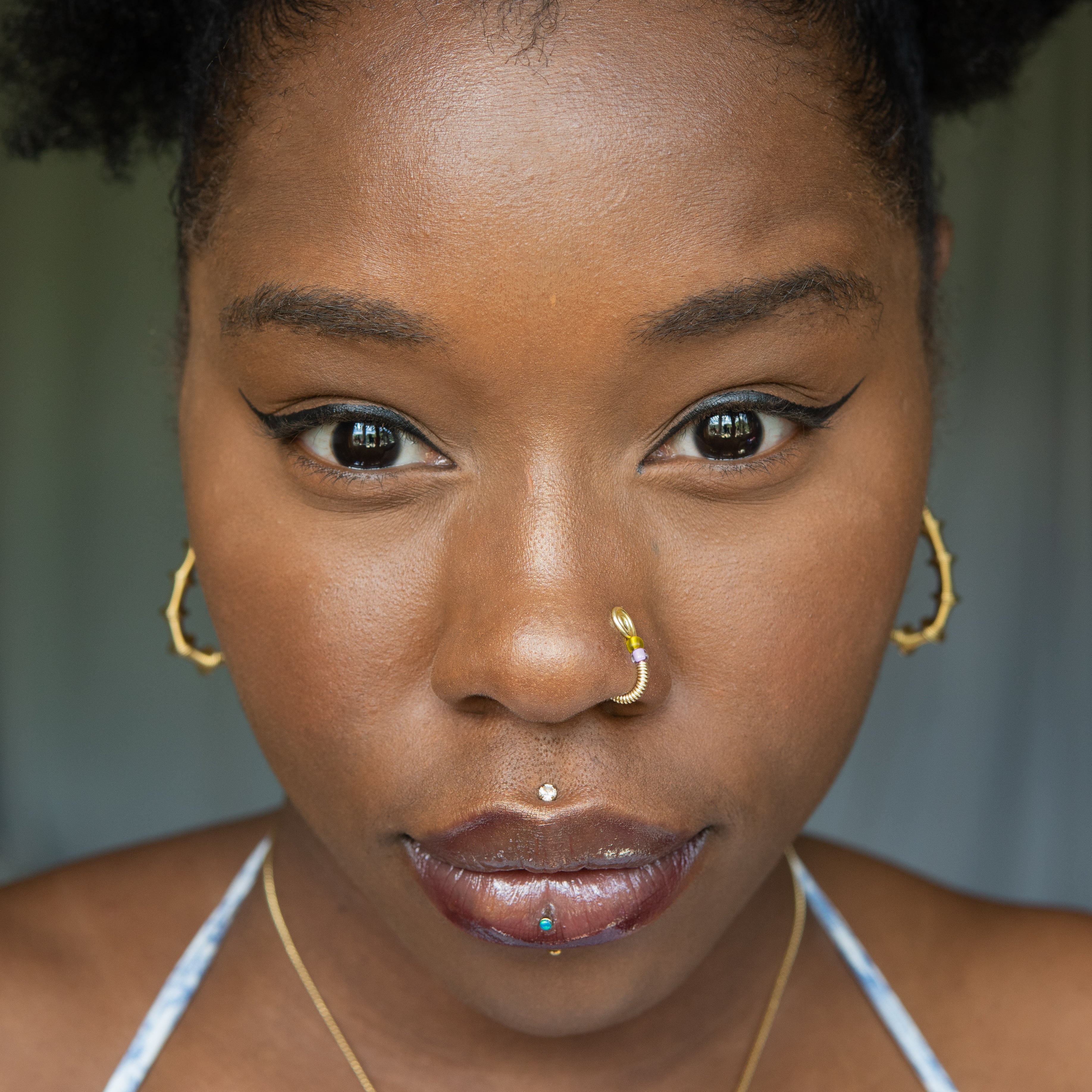 A light gold-toned fake nose ring cuffs the model's nostril. The wire design forms a circular shape. Two seed beads (yellow and lilac) rest below the circle.