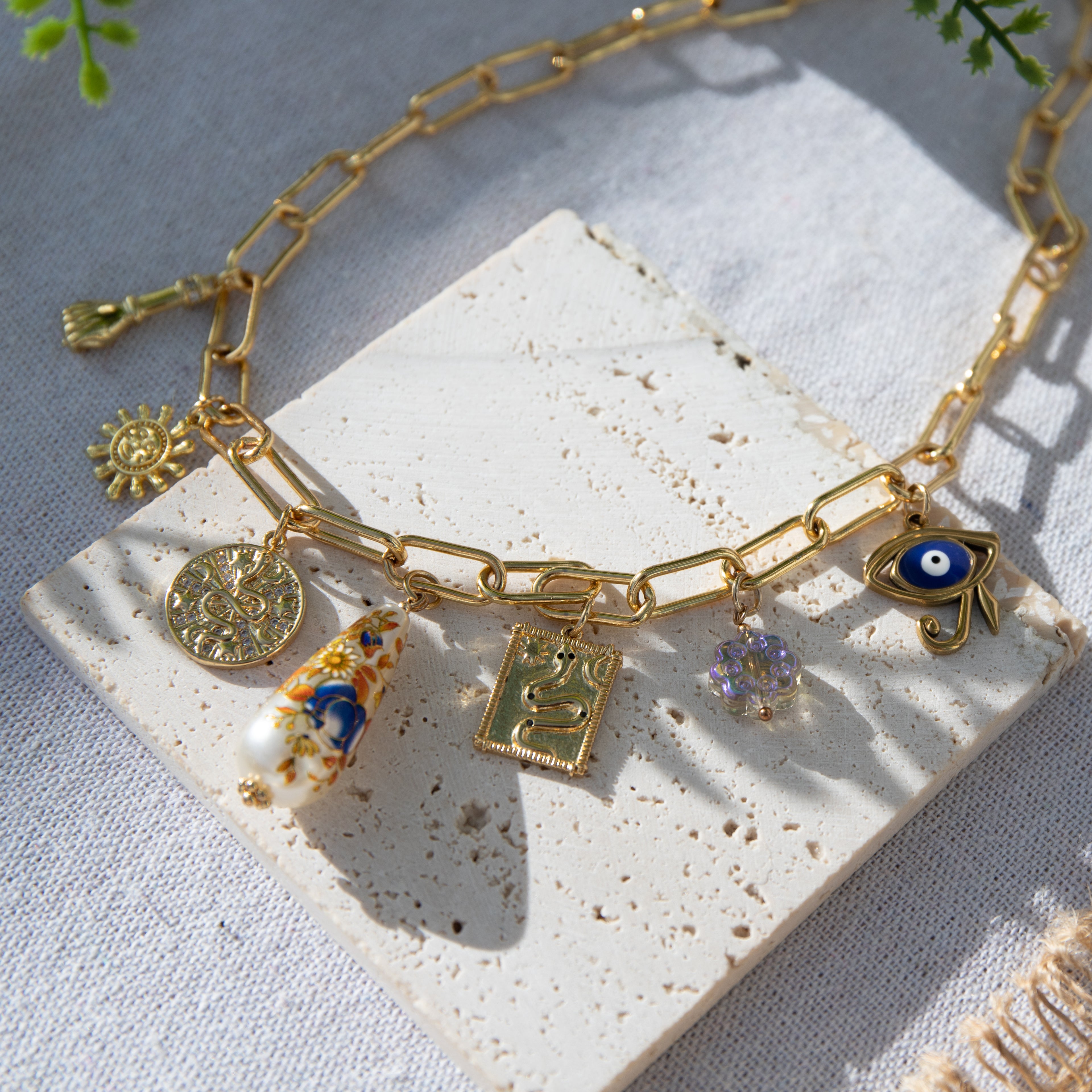 A chunky gold-plated charm necklace made with a chunky 18K gold-plated curb chain. Multiple gold-plated charms hang along the length of the necklace in the following order: seashell, padlock, scarab beetle, a green heart, crescent moon, Duafe symbol, circle disc with the engravement 'thankful, grateful, blessed'