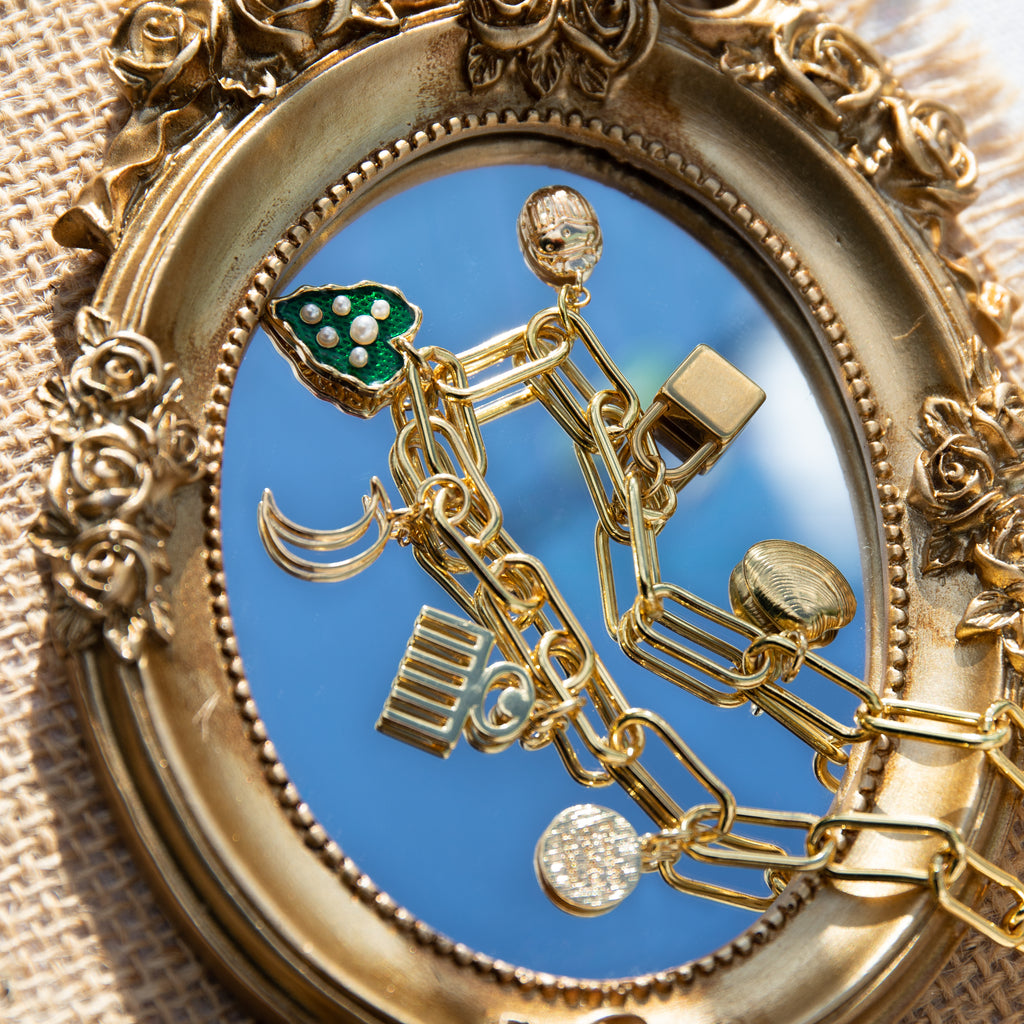 A chunky gold-plated charm necklace made with a chunky 18K gold-plated curb chain. Multiple gold-plated charms hang along the length of the necklace in the following order: seashell, padlock, scarab beetle, a green heart, crescent moon, Duafe symbol, circle disc with the engravement 'thankful, grateful, blessed'