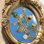 A chunky gold-plated charm necklace made with a chunky 18K gold-plated curb chain. Multiple gold-plated charms hang along the length of the necklace in the following order: seashell, padlock, scarab beetle, a green heart, crescent moon, Duafe symbol, circle disc with the engravement 'thankful, grateful, blessed'