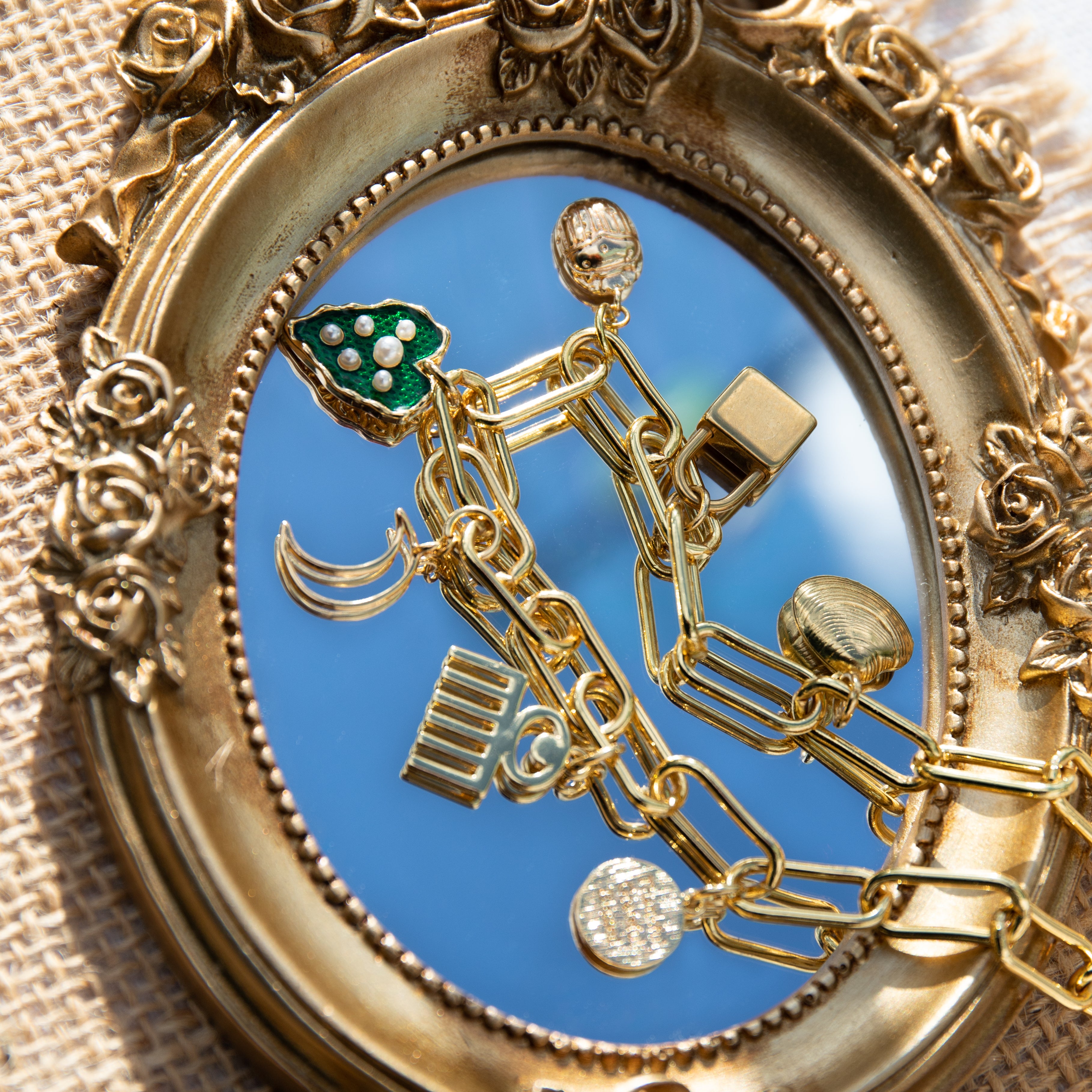 A chunky gold-plated charm necklace made with a chunky 18K gold-plated curb chain. Multiple gold-plated charms hang along the length of the necklace in the following order: seashell, padlock, scarab beetle, a green heart, crescent moon, Duafe symbol, circle disc with the engravement 'thankful, grateful, blessed'