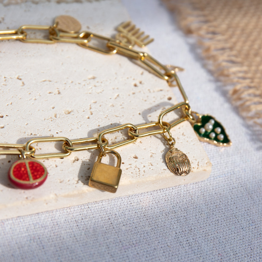 A chunky gold-plated charm necklace made with a chunky 18K gold-plated curb chain. Multiple gold-plated charms hang along the length of the necklace in the following order: pomegranate, padlock, scarab beetle, a green heart, crescent moon, Duafe symbol, circle disc with the engravement 'thankful, grateful, blessed'