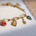 A chunky gold-plated charm necklace made with a chunky 18K gold-plated curb chain. Multiple gold-plated charms hang along the length of the necklace in the following order: pomegranate, padlock, scarab beetle, a green heart, crescent moon, Duafe symbol, circle disc with the engravement 'thankful, grateful, blessed'