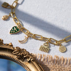 A chunky gold-plated charm necklace made with a chunky 18K gold-plated curb chain. Multiple gold-plated charms hang along the length of the necklace in the following order: seashell, padlock, scarab beetle, a green heart, crescent moon, Duafe symbol, circle disc with the engravement 'thankful, grateful, blessed'