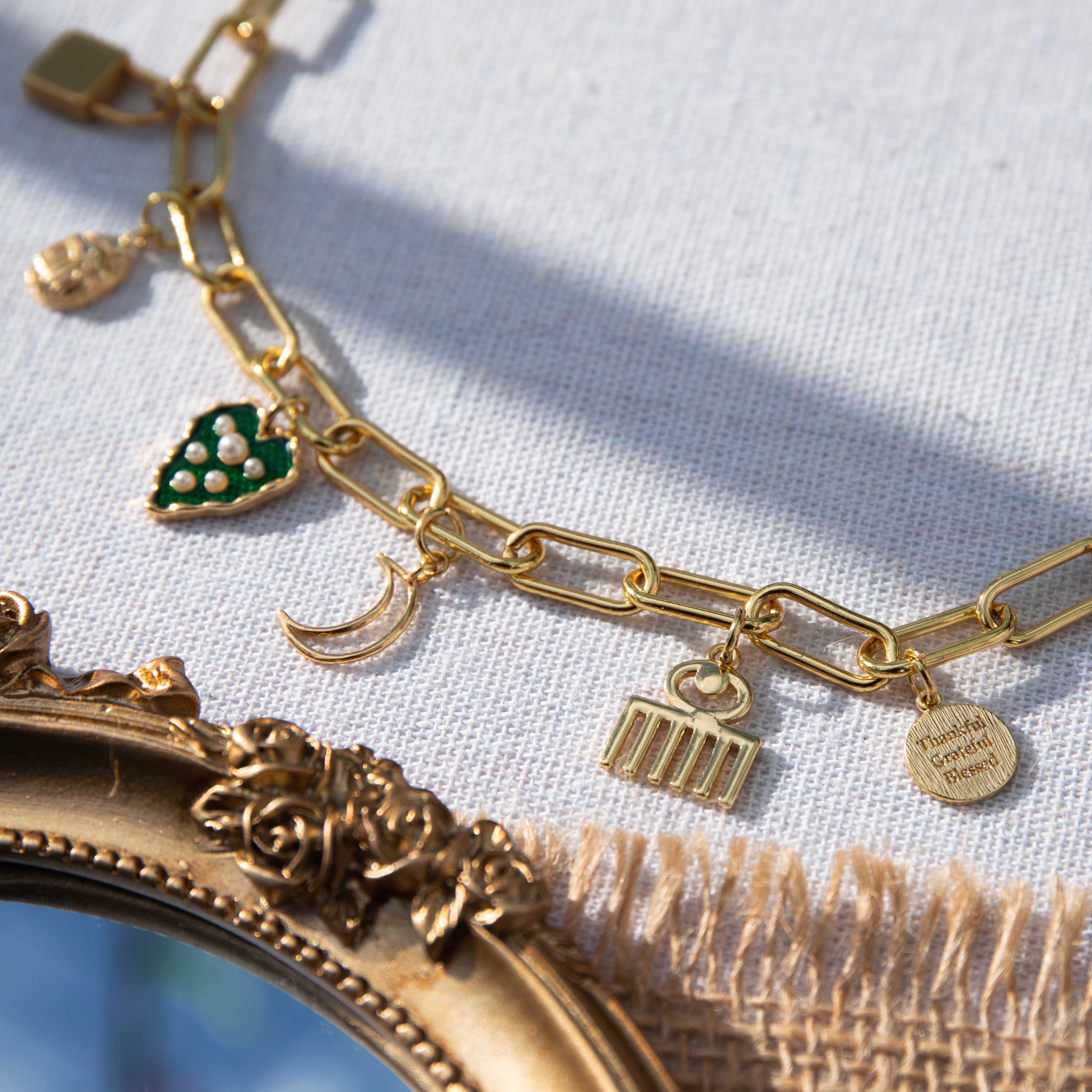 A chunky gold-plated charm necklace made with a chunky 18K gold-plated curb chain. Multiple gold-plated charms hang along the length of the necklace in the following order: seashell, padlock, scarab beetle, a green heart, crescent moon, Duafe symbol, circle disc with the engravement 'thankful, grateful, blessed'