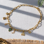 A chunky gold-plated charm necklace made with a chunky 18K gold-plated curb chain. Multiple gold-plated charms hang along the length of the necklace in the following order: seashell, padlock, scarab beetle, a green heart, crescent moon, Duafe symbol, circle disc with the engravement 'thankful, grateful, blessed'