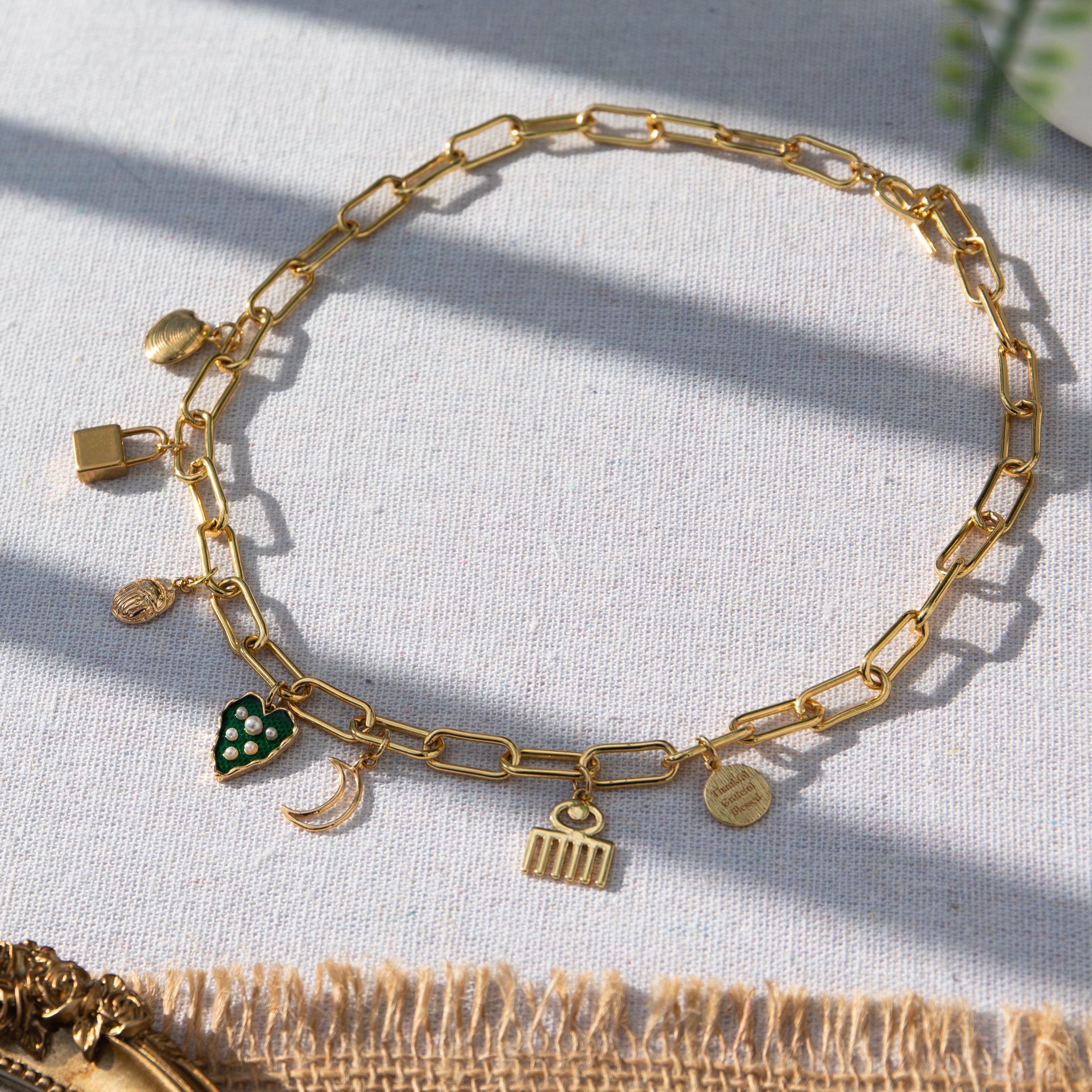 A chunky gold-plated charm necklace made with a chunky 18K gold-plated curb chain. Multiple gold-plated charms hang along the length of the necklace in the following order: seashell, padlock, scarab beetle, a green heart, crescent moon, Duafe symbol, circle disc with the engravement 'thankful, grateful, blessed'