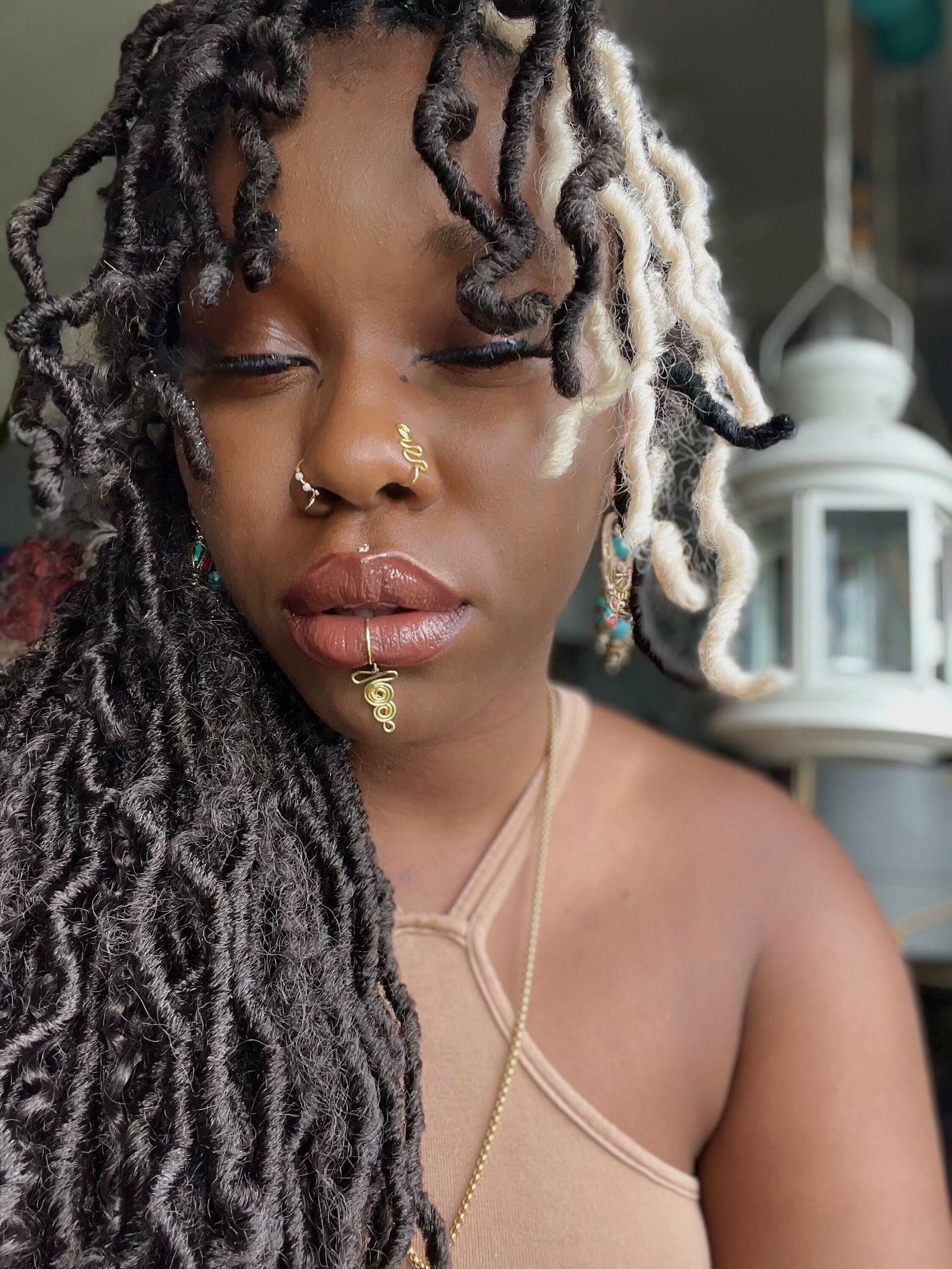 A gold-toned, tightly spiralled nose cuff sits on the model's nose. The design begins with a tightly formed circle that gradually twists in a wavy pattern. It is accompanied by a faux lip ring cuffing the model's bottom lip. The flexible wire is tightly moulded to create a circular spiral shape that gradually decreases in size.