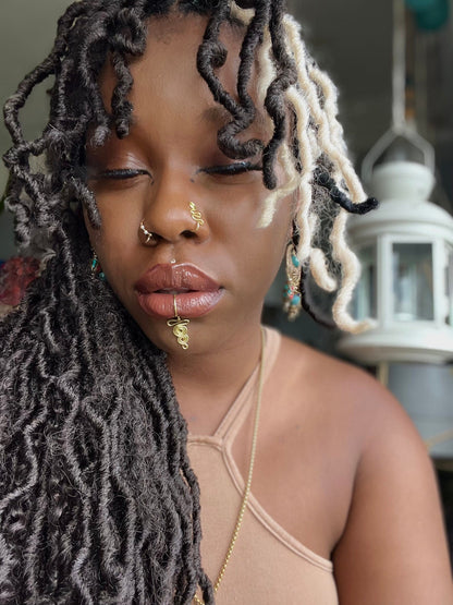 A gold-toned, tightly spiralled nose cuff sits on the model's nose. The design begins with a tightly formed circle that gradually twists in a wavy pattern. It is accompanied by a faux lip ring cuffing the model's bottom lip. The flexible wire is tightly moulded to create a circular spiral shape that gradually decreases in size.